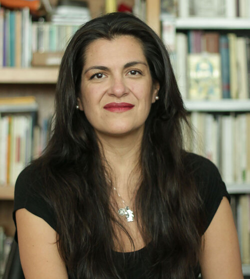 Woman with long brown hair in front of blurred books on a bookcase.