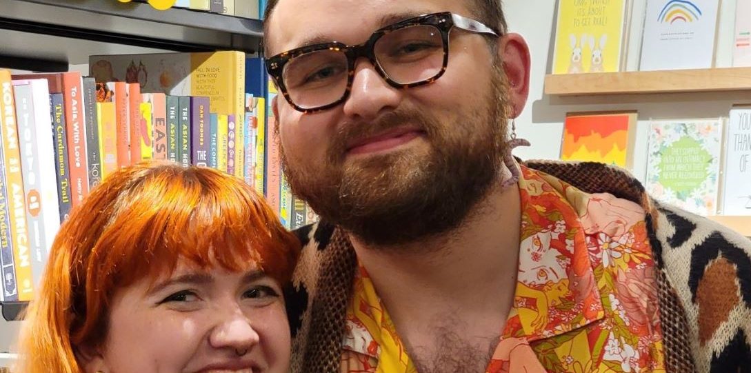 Man with glasses and woman with red hair pose together in front of bookshelves.