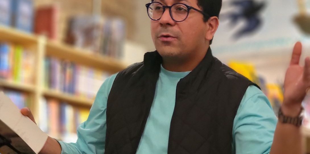 Man with short hair and glasses in a long sleeve teal shirt and black vest. He is holding a book and reading from it.