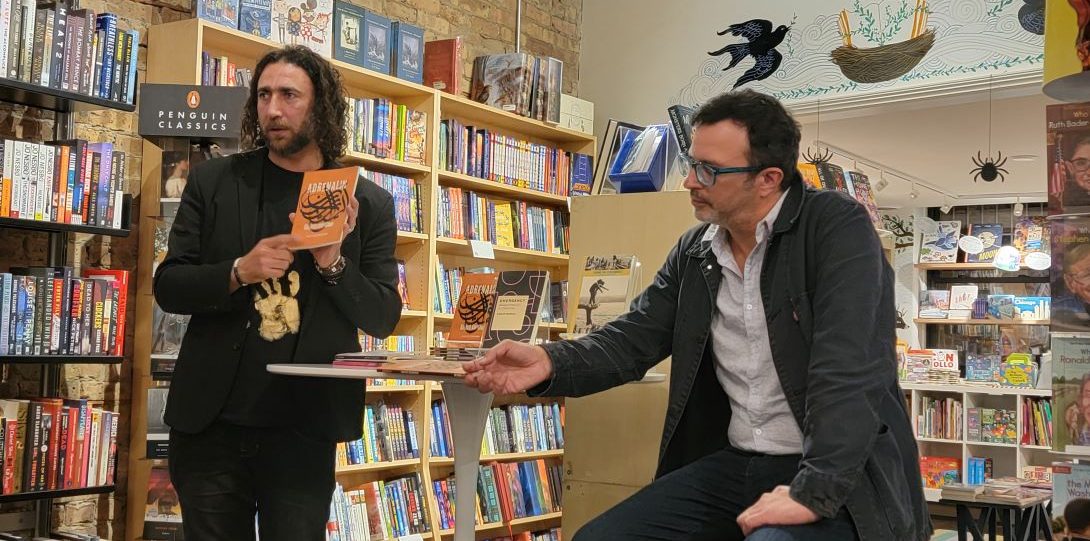 Man with long curly hair and light skin holds up a book while standing. Man with short hair and glasses sits on stool as he listens to the other man. Both are in a bookstore.