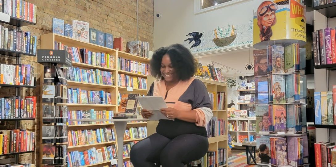 Young black woman sitting on a stool reading from her paper. She is sitting in front of a bookcase.