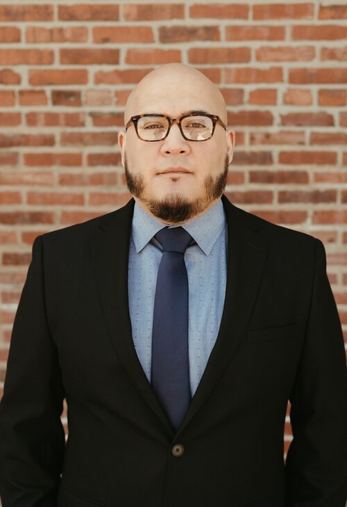 Man in a suit with glasses and a beard standing in front of a brick wall.
