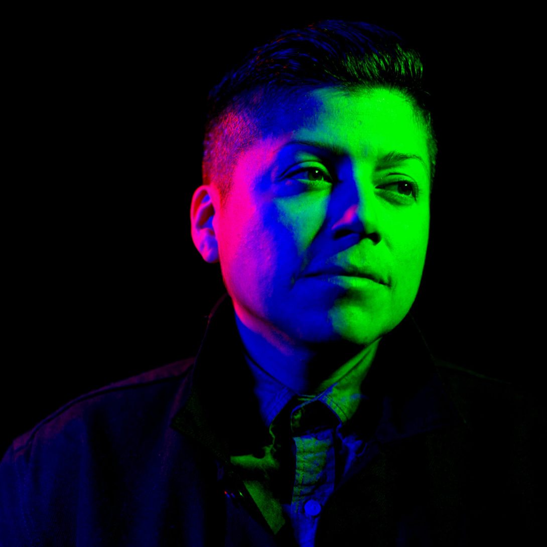 Author portrait with green, blue, purple, and pink lights shining on their face among a black background.