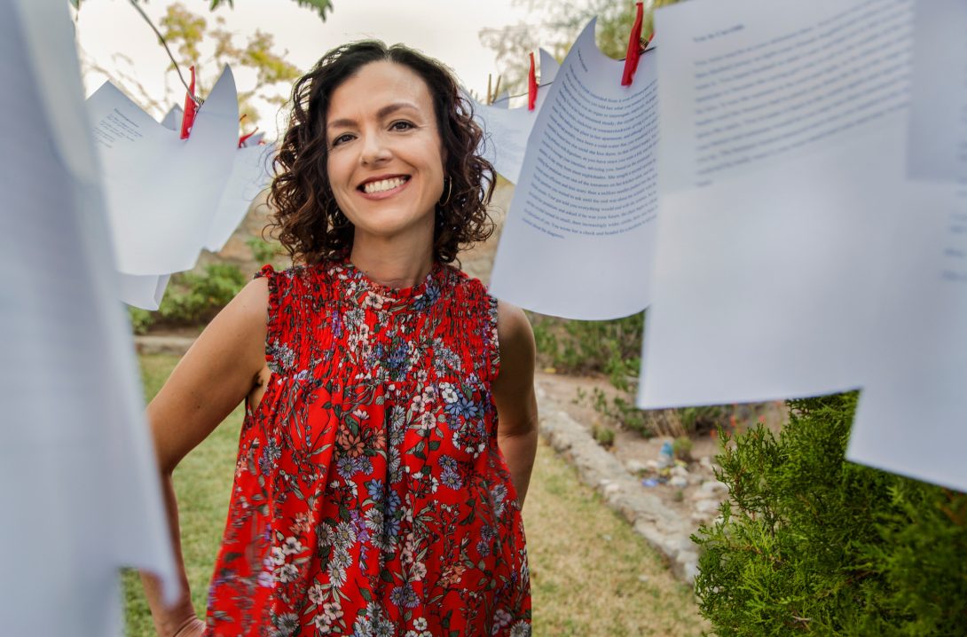 Woman with short curly hair in a red shirt. Pages with blurred written text surround her, hung up by clothes pins.